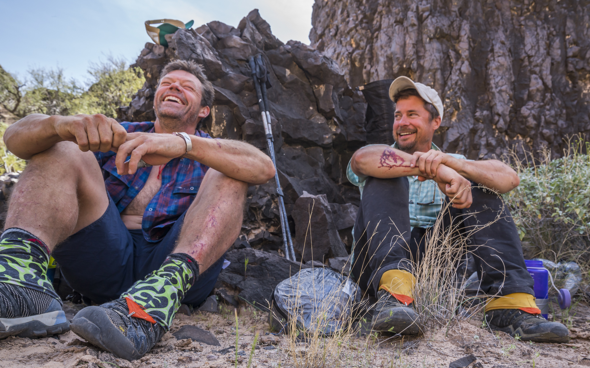 Kevin Fedarko and Pete McBride sit against a rock. They are outfitted in hiking shoes. They laugh.