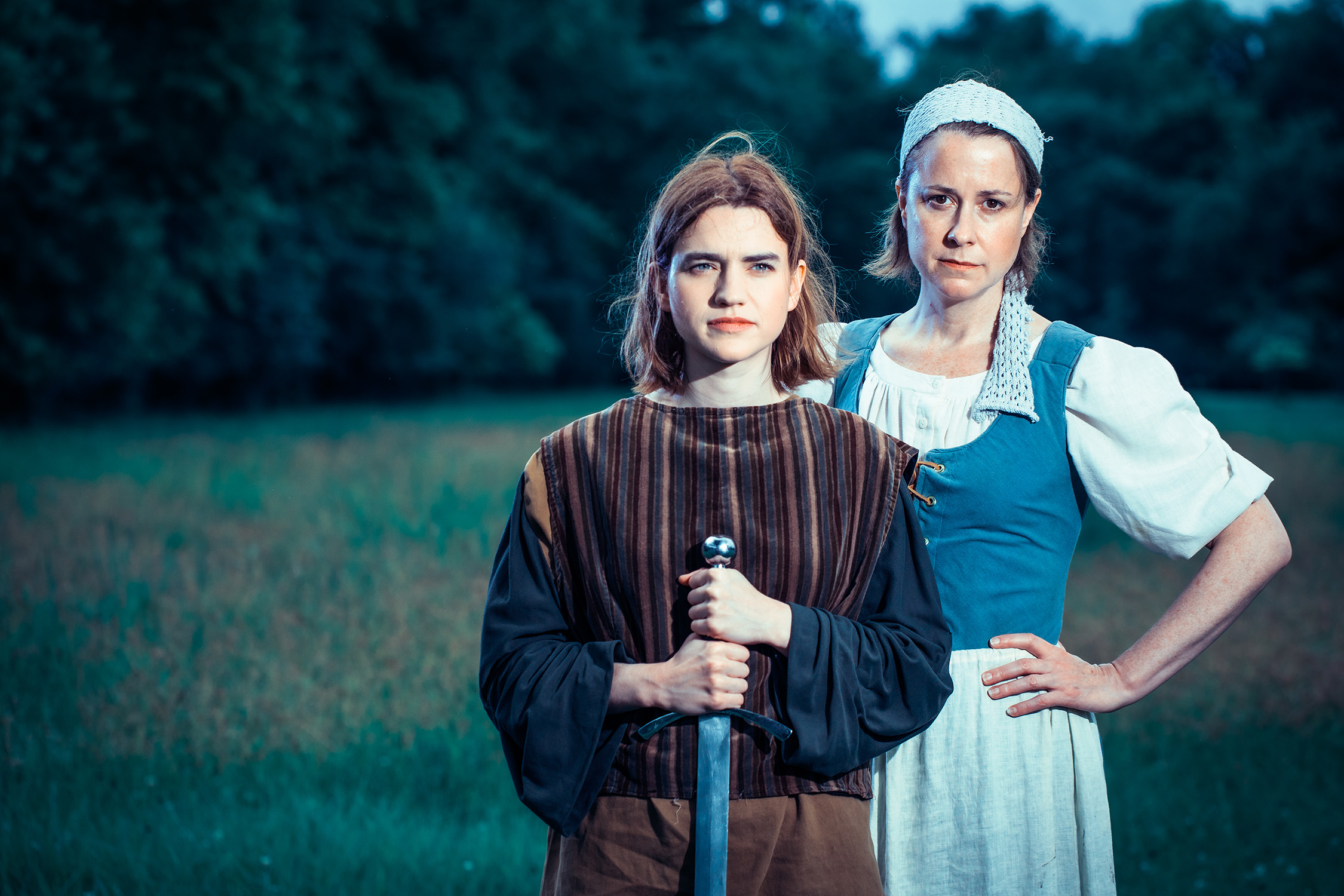 Two people stand in a field, face forward. One wears a brown tunic and clasps a sword, pointed downward with both hands. The other wears a blue maiden attire, one hand on their hip.
