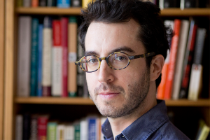A person wearing glasses stands in front of a bookshelf and gazes at the viewer.